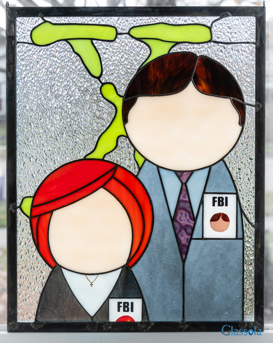 stained glass panel featuring a minimalist design of Mulder and Scully from The X-Files standing in front of the iconic "X" logo. The panel is shown illuminated from behind by window light.