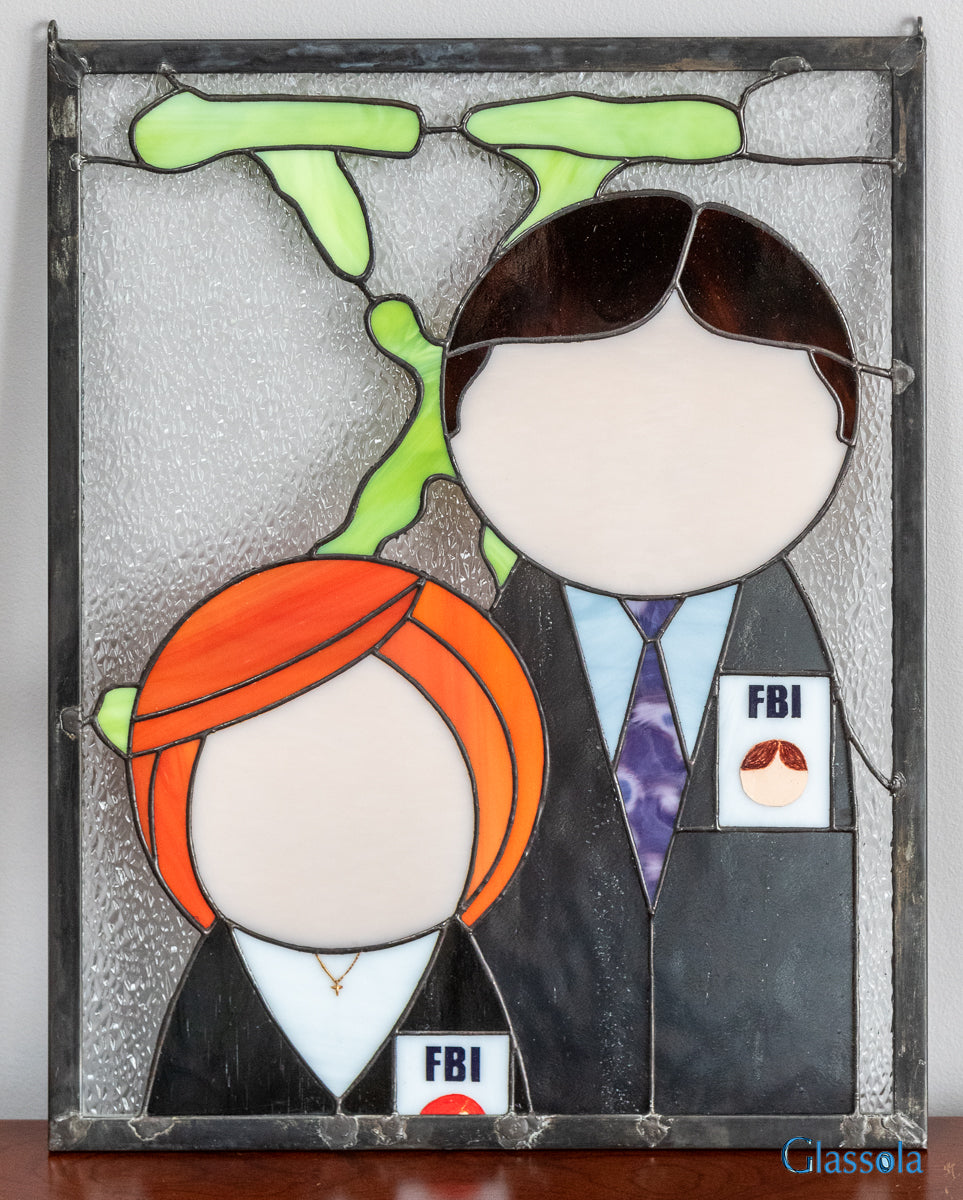 stained glass panel featuring a minimalist design of Mulder and Scully from The X-Files standing in front of the iconic "X" logo. The panel is shown in ambient room light against a wall.